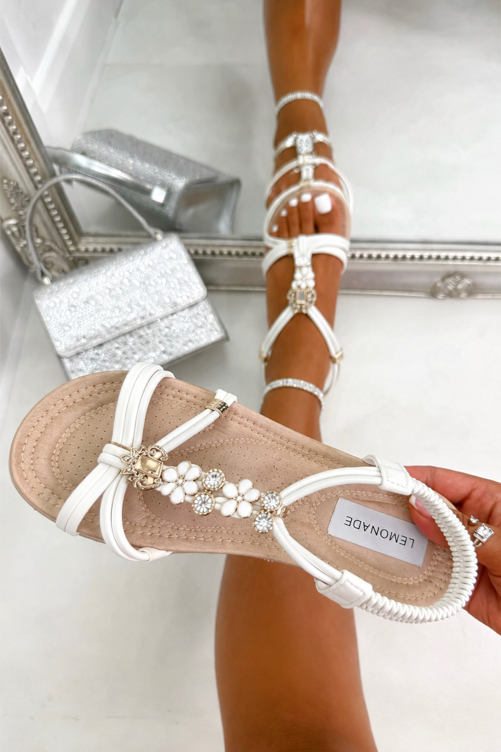 White strappy flat sandals with pearl and crystal embellishments on the toe strap, shown worn on a foot and held in hand against a light tiled background with a mirror reflection.