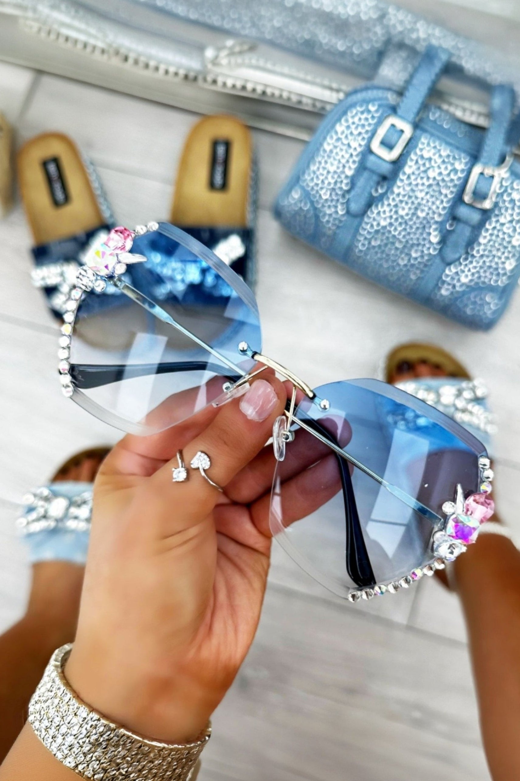 Blue embellished sunglasses held by a hand with a glittery bracelet, with shoes and a bag in the background.