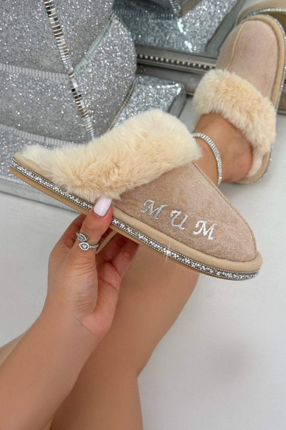 Beige slippers with fur lining held by a hand, with a sparkly silver prop in the background.