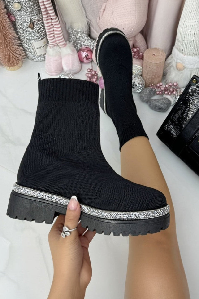 Black platform boots with rhinestone trim held by a hand, with a background of colorful yarn and a black bag.