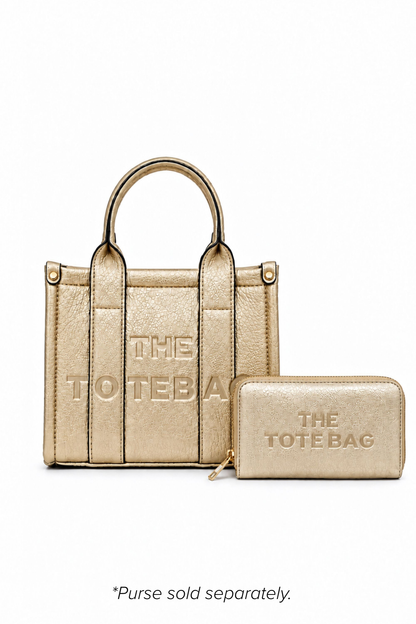 Champagne Gold mini Tote bag with with two rounded top handles, embosses "THE TOTE BAG" lettering on the front, vertical stitched panels, gold-tone stud fastenings and a small side tab detail. Pictured with champagne tote purse in front.