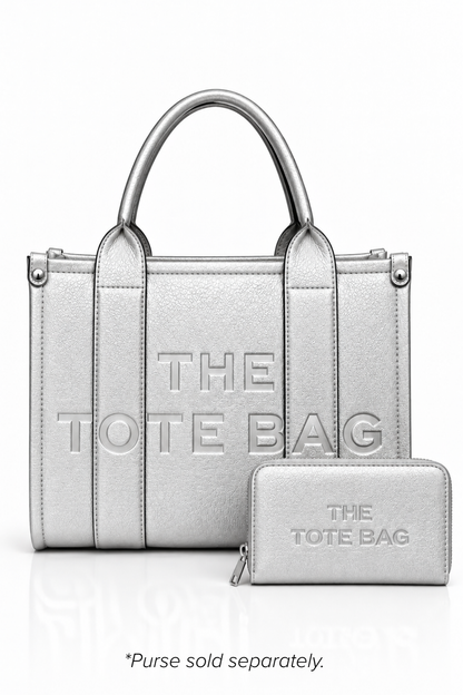 Metallic Silver structured tote bag with two rounded top handles, embossed. "THE TOTE BAG" lettering on the front, vertical stitched panels, contrast edging and gold- tone stud details.  Metallic Silver Tote purse pictured in front. Set against a white background.