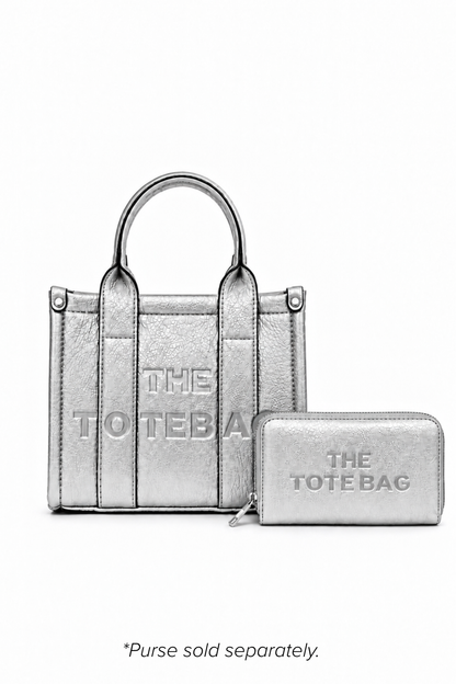 Silver mini structured tote bag with two rounded top handles, vertical stitched panels, matching tonal stitching and subtle embossed "THE TOTE BAG" lettering on the front. Shown with matching decorative key ring and rectangular zip purse. Purse and keyring sold separately.  