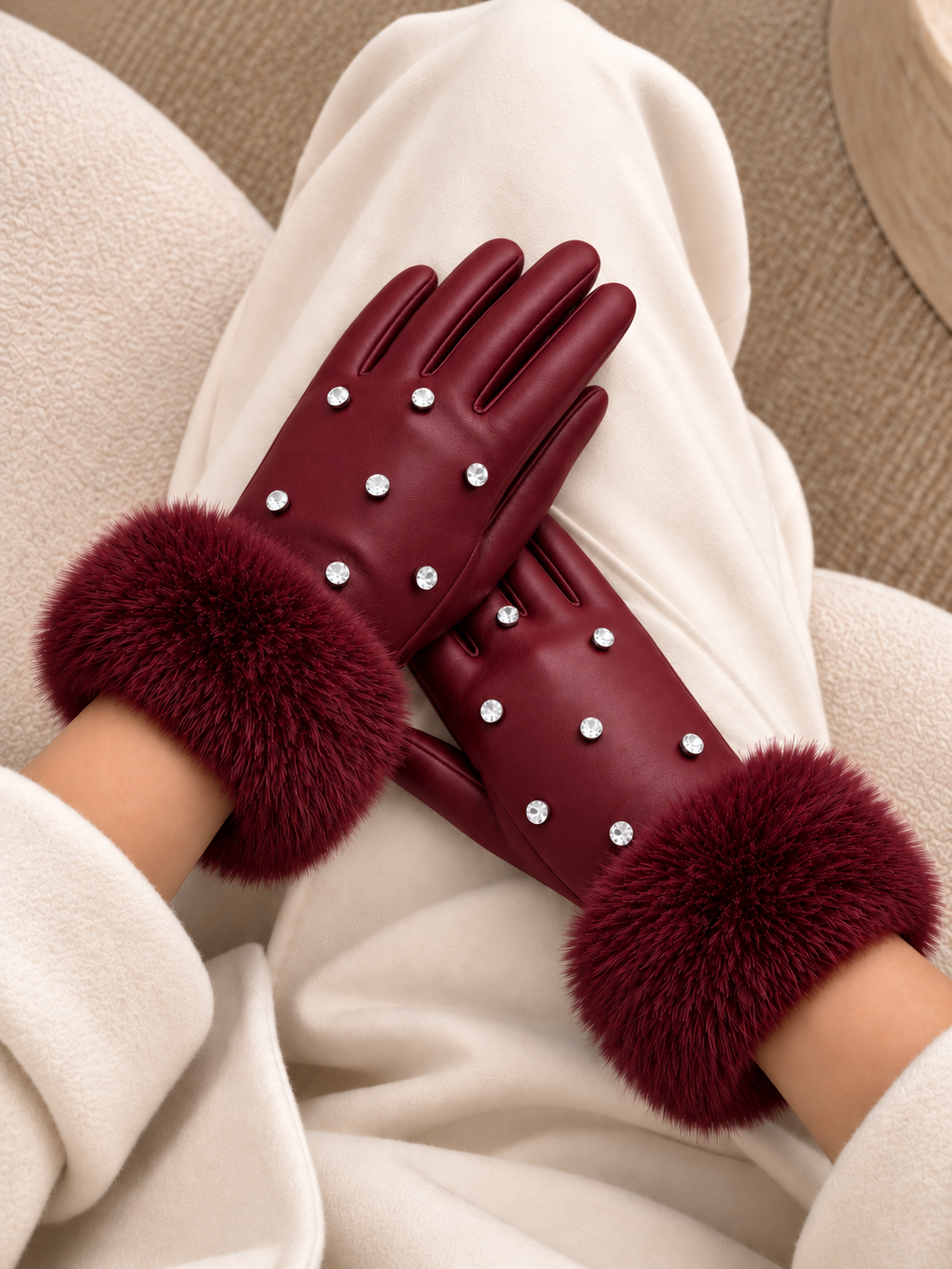 A pair of burgundy leather gloves with fluffy faux-fur cuffs and small rhinestone embellishments, worn on hands resting on cream-colored clothing.