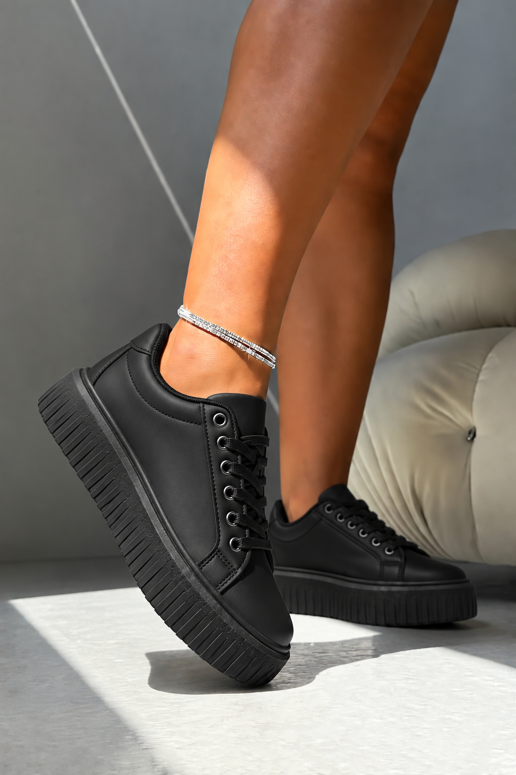 A person wearing black low-top platform sneakers with chunky lug soles and black laces, shown from the knees down in a modern indoor setting with a gray wall and light-colored cushion.