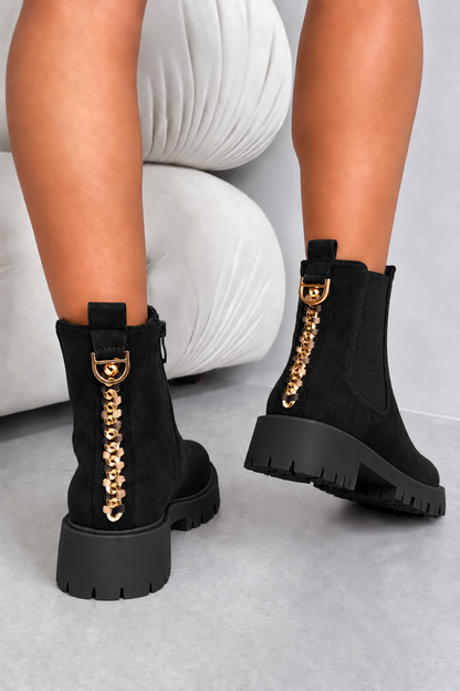 Black ankle boots with chunky lug soles and a prominent gold-tone chain detail with a horsebit hardware accent, shown on a model&