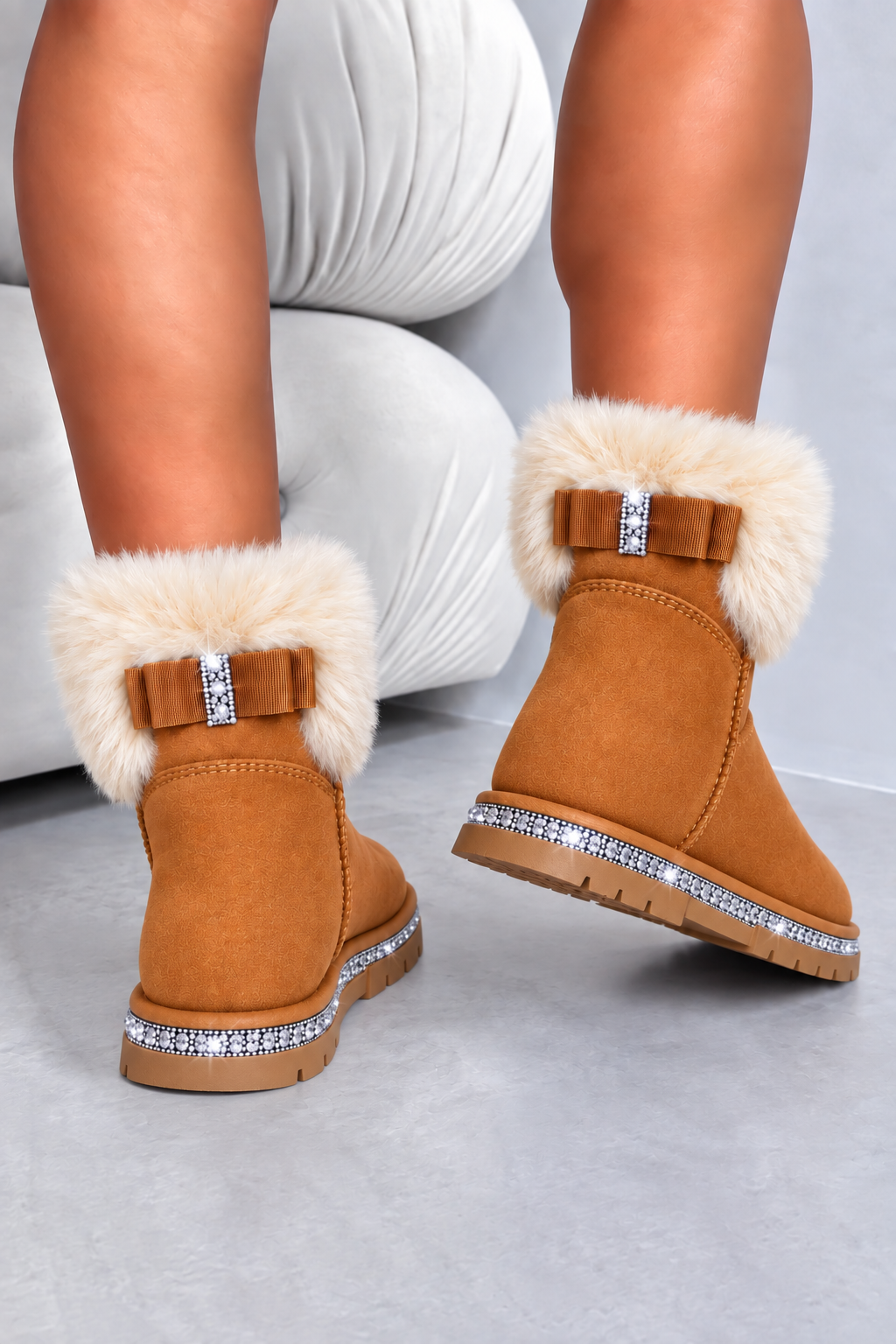 A pair of tan suede ankle boots with fluffy cream faux-fur trim, decorative bow details with rhinestone accents, and crystal-studded soles, shown on a model&