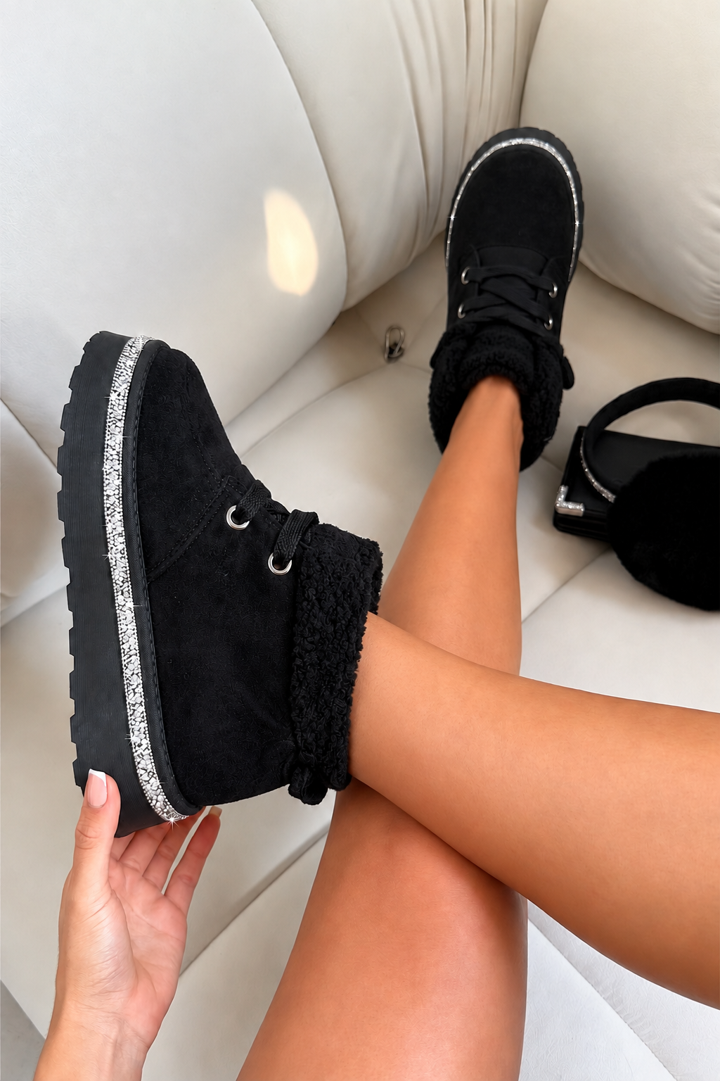 A person wearing black suede ankle boots with black faux-fur trim and a silver glitter sole, sitting on a white leather couch. One boot is being held up by a hand, and a black headband is visible on the couch.