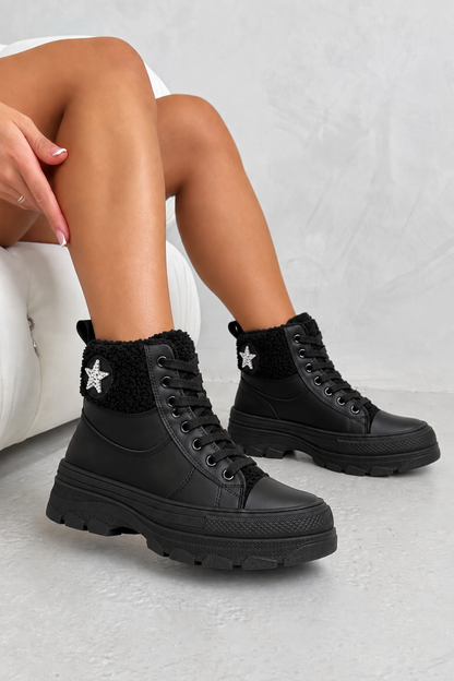 A person wearing black high-top winter boots with black faux-fur trim and a silver star detail on the ankle, seated against a light gray textured background.