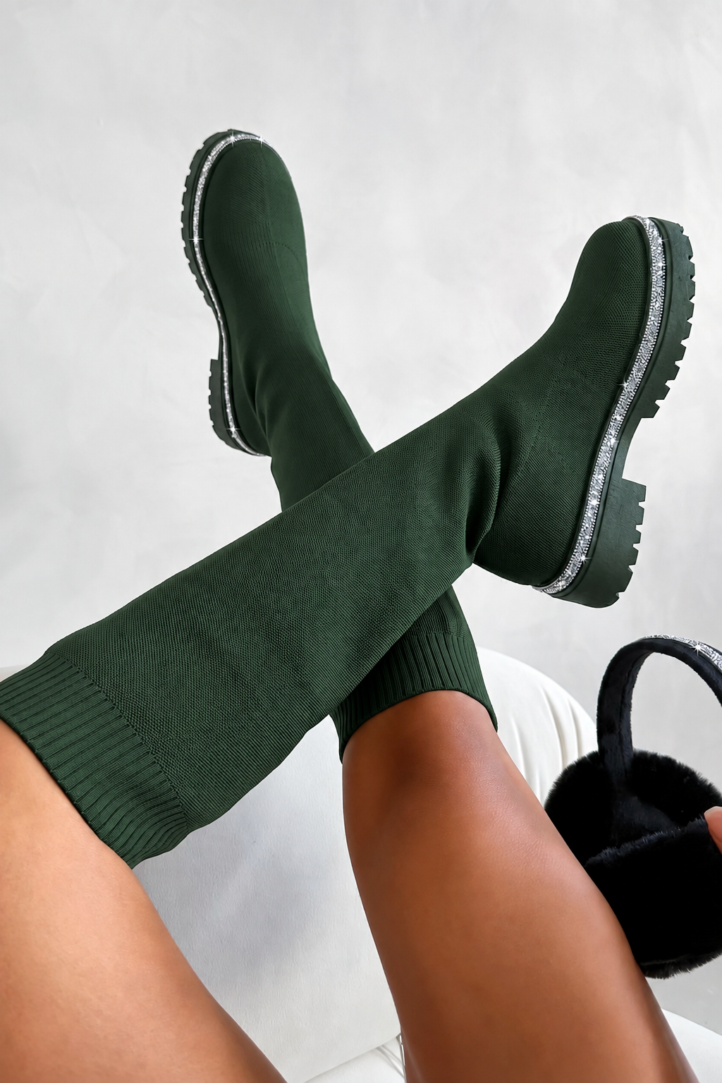 A person wearing dark forest green knit boot covers with chunky black soles, crossed at the ankles, with a black furry slippers visible in one hand against a light gray background.