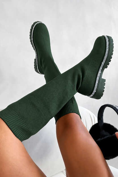 A person wearing dark forest green knit boot covers with chunky black soles, crossed at the ankles, with a black furry slippers visible in one hand against a light gray background.