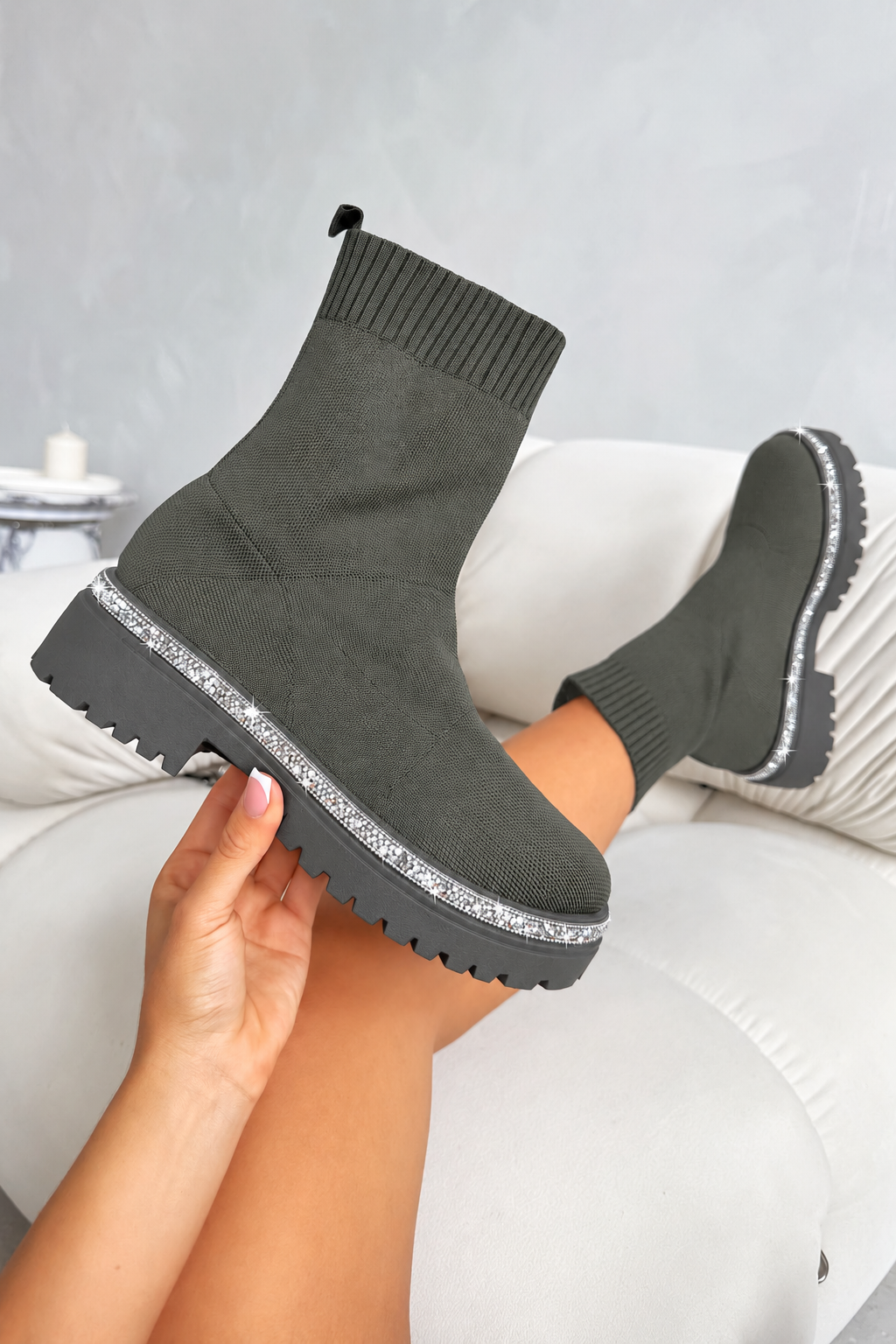 A pair of dark olive green sock-style ankle boots with chunky lug soles and a silver rhinestone trim, shown being worn with one boot lifted by a hand in front of a white sofa.