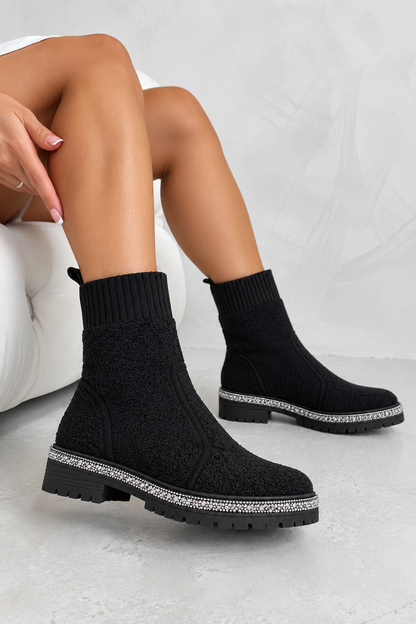 A person wearing black knit ankle boots with chunky lug soles and a silver rhinestone trim, seated on a white tufted chair against a light gray background.