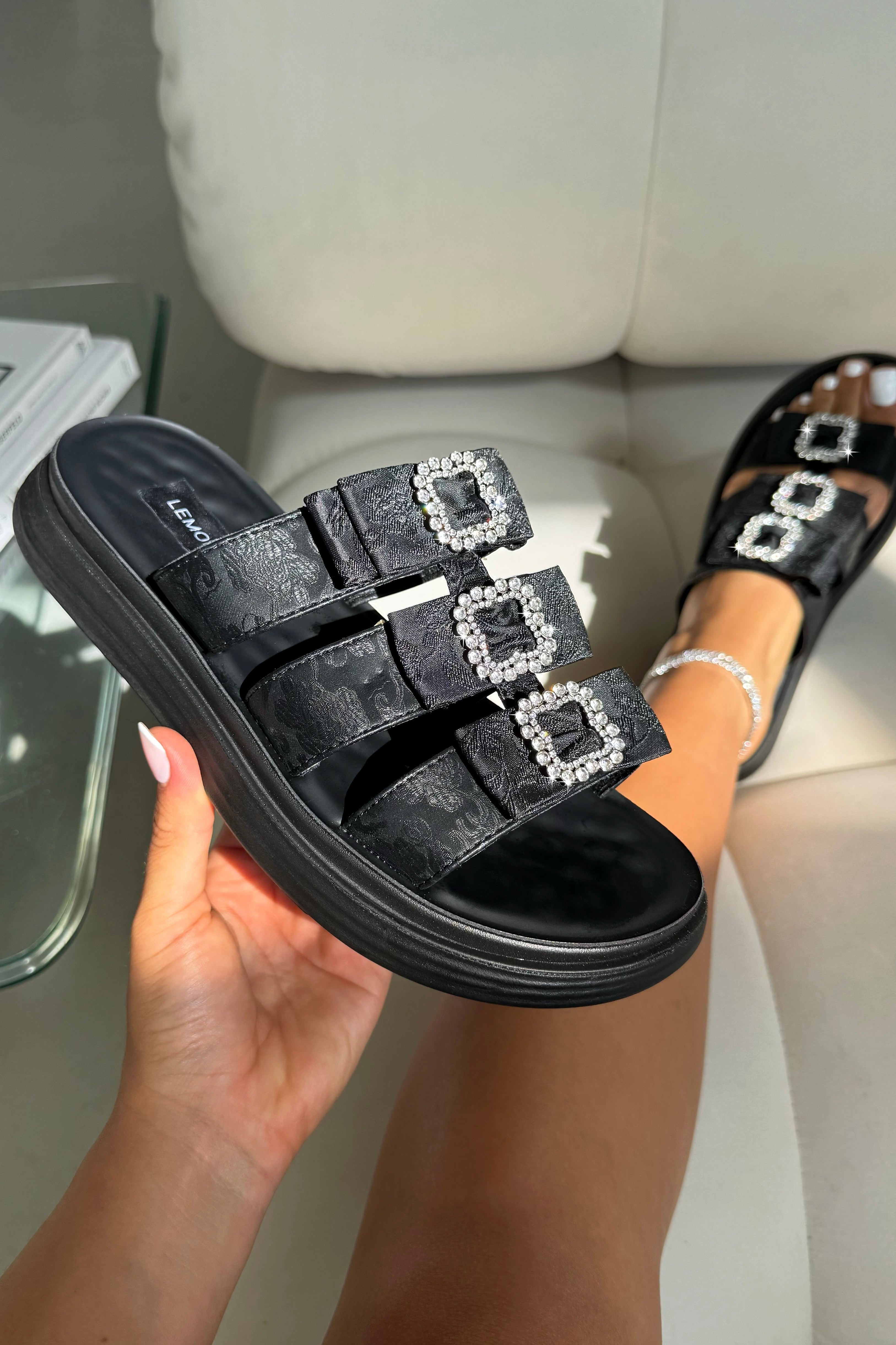 Black slide sandals with embellishments held by a hand on a light-colored surface.