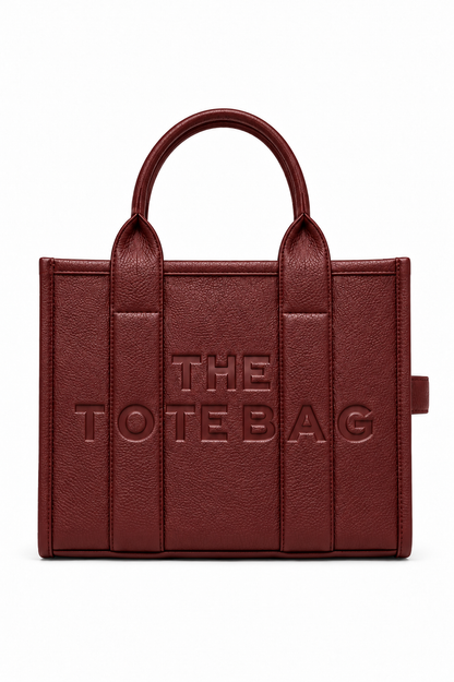 Burgundy Faux leather tote bag with two rounded top handles, embossed "THE TOTE BAG" lettering on the front, contrast stitching. gold tone hardware. Set against a white background.