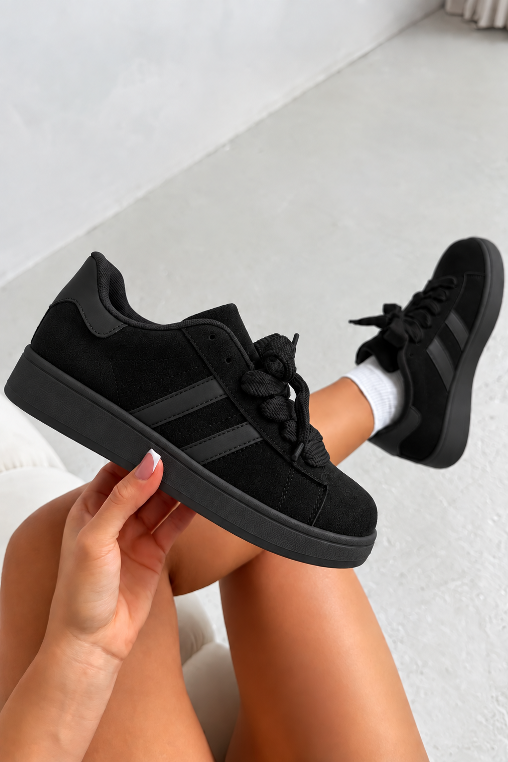 A pair of black low-top sneakers with textured laces and three dark stripes, shown being held by a person&