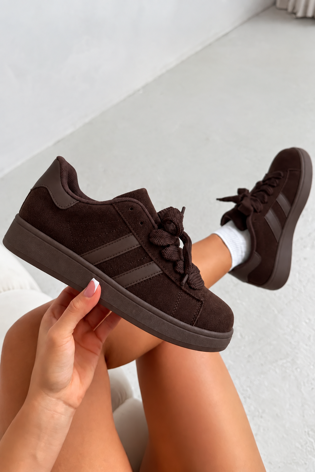 A pair of dark brown suede low-top sneakers with matching dark brown laces and side stripes, shown being worn with white socks, held by a person&