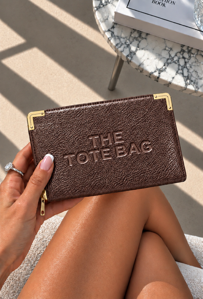 A person holds a brown textured zip-around wallet or clutch with gold-tone corner hardware and embossed &