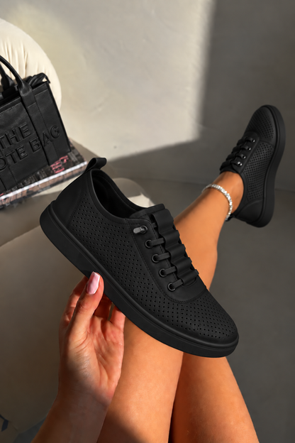 A person holds up a black perforated sneaker while sitting in a chair, with the other shoe worn on their foot. A black tote bag is visible in the background.