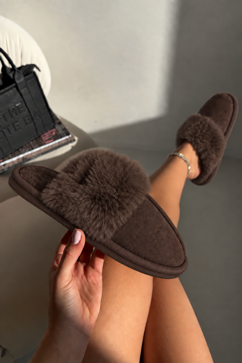 A person holds a brown fuzzy slide slipper in one hand while wearing the same slipper on their foot, with a gray wall and a black handbag in the background.