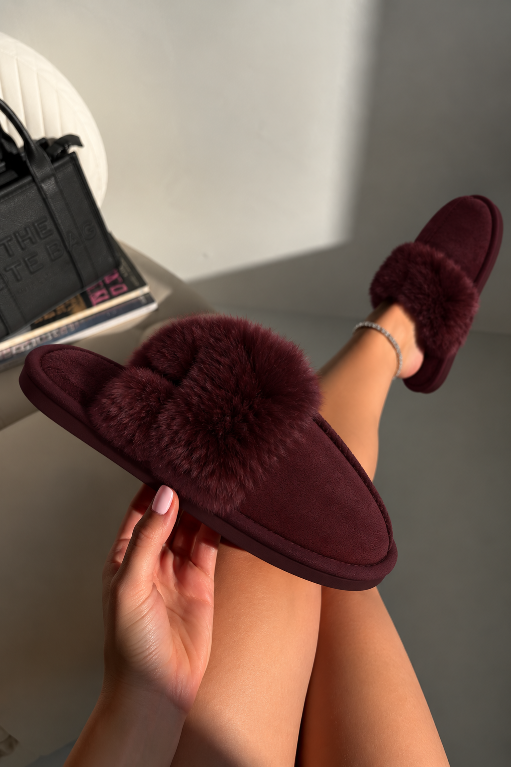 A person holds a pair of deep burgundy slippers with fluffy faux-fur trim in one hand while wearing the matching slippers on their feet, set against a neutral indoor background.