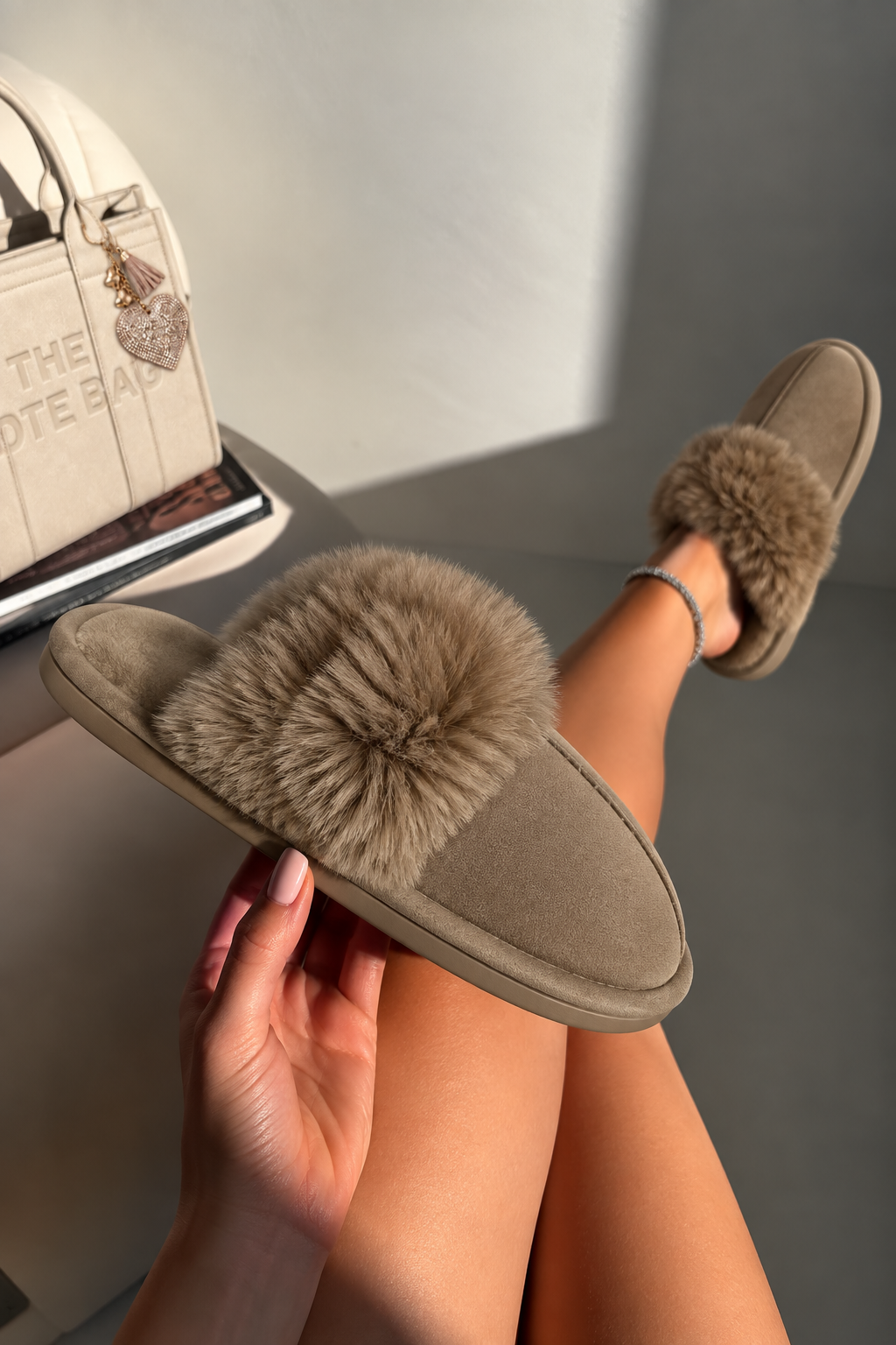 A person holds a taupe suede and faux fur slipper in one hand while wearing the matching slipper on their foot, with a cream handbag visible in the background.