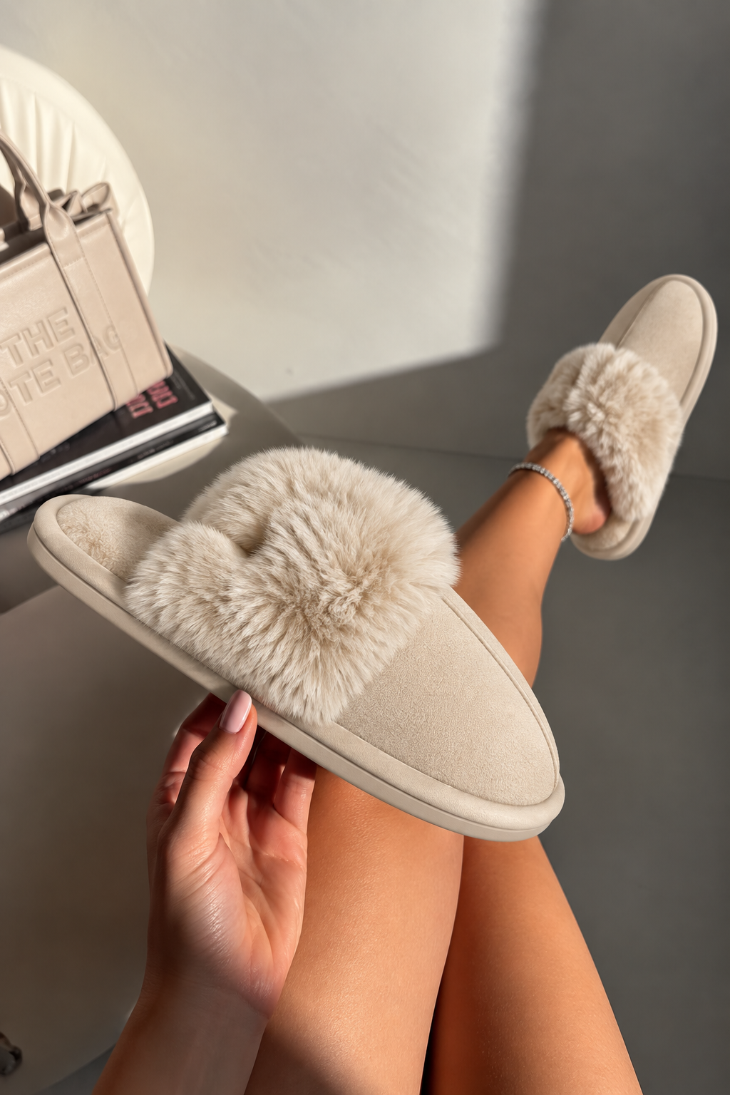 A person wearing beige slip-on slippers with fluffy cream faux-fur trim, shown in a styled indoor setting with one slipper held in hand and the other on the foot.