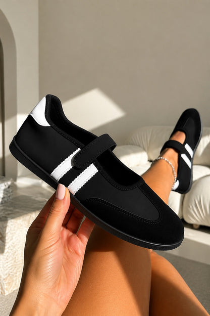 A person holds a black and white low-top shoe in one hand while wearing the same shoe on the other foot in a bright indoor setting with a white sofa in the background.
