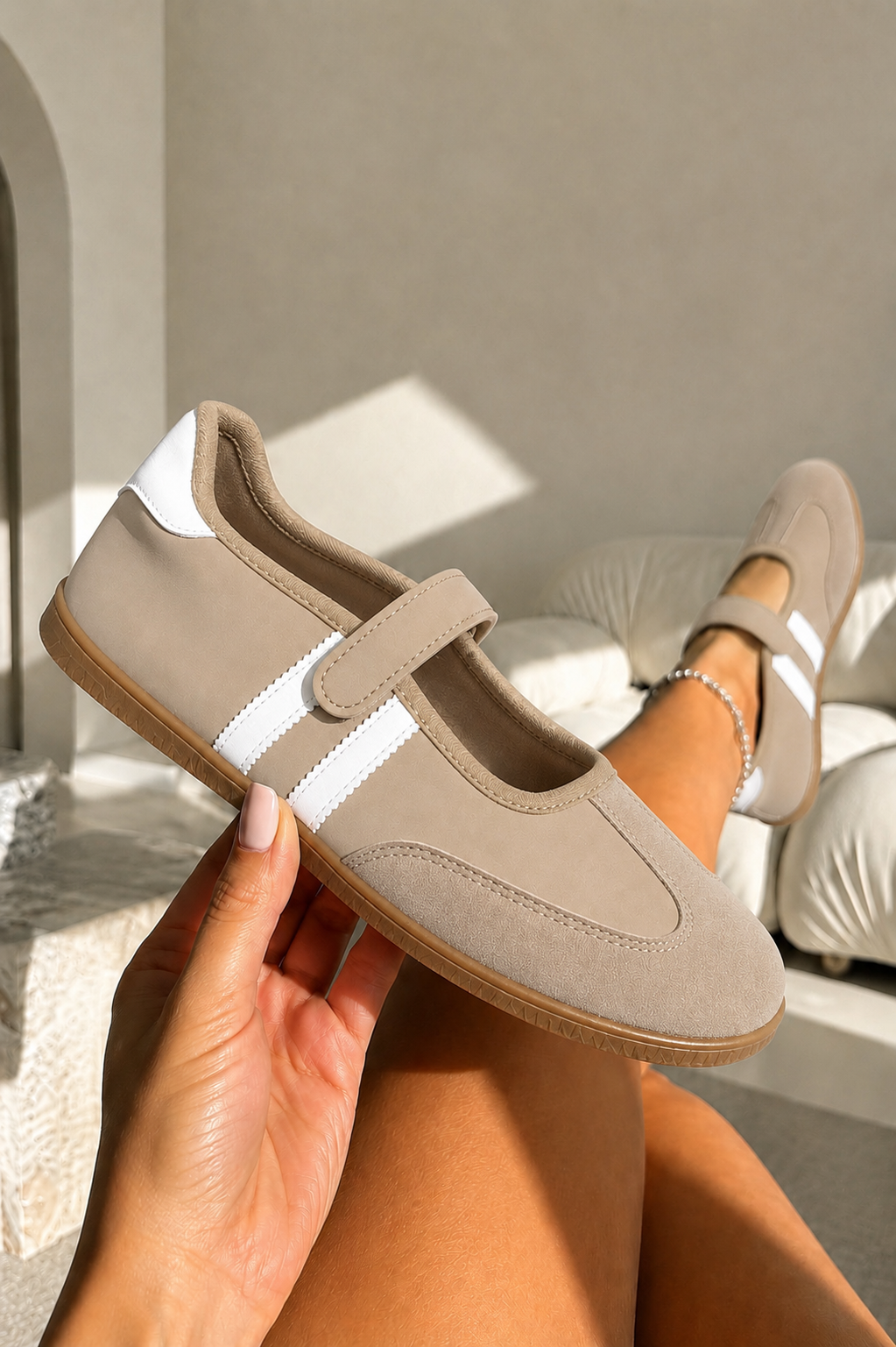 A person holds a taupe faux suede Mary Jane shoe in one hand while wearing the same shoe on their foot, set in a bright, modern room with warm sunlight and neutral walls.
