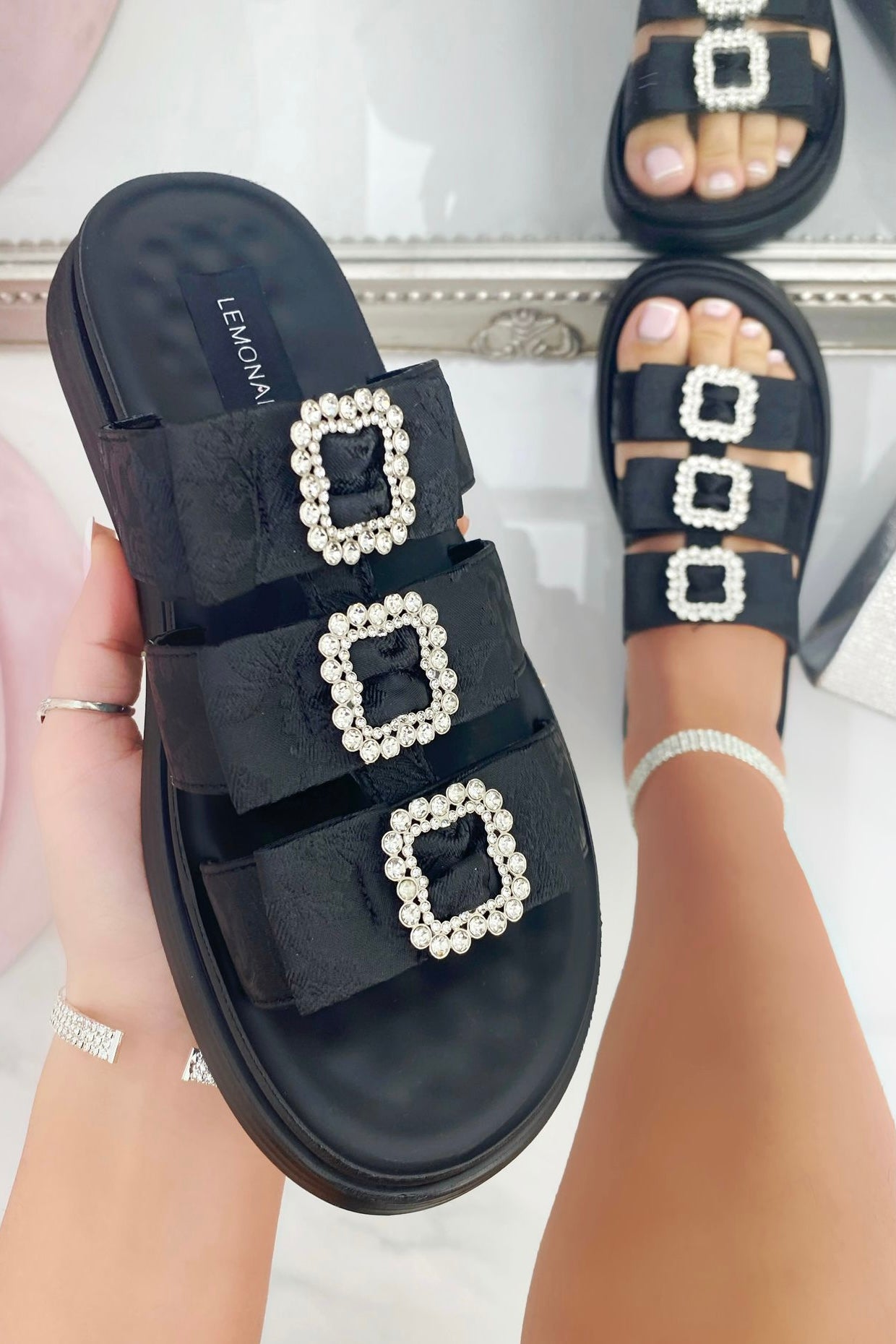 Black sandals with square pearl embellishments worn by a person on a light background