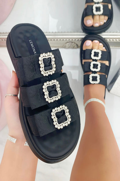 Black sandals with square pearl embellishments worn by a person on a light background