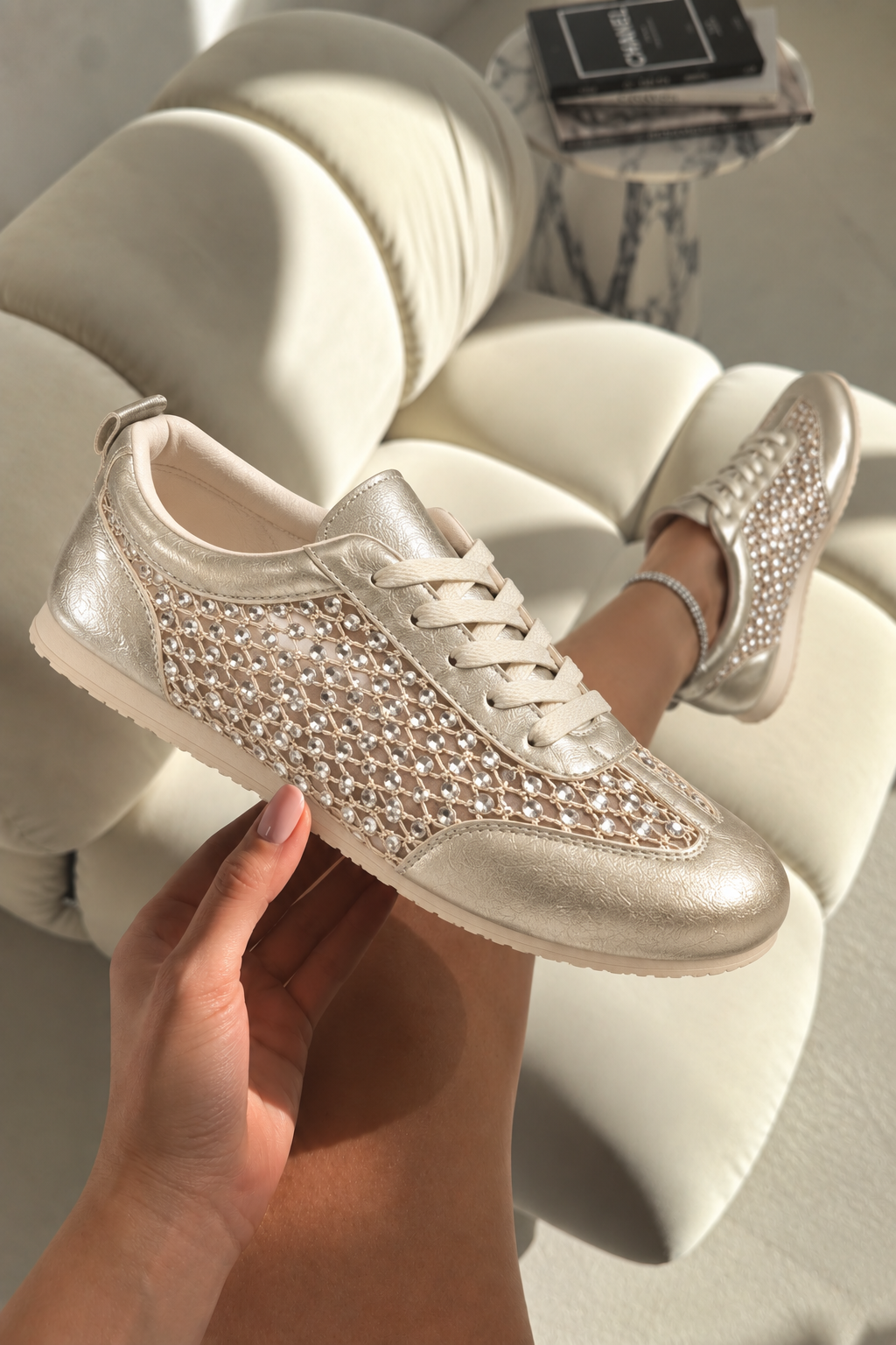 A pair of metallic champagne-gold sneakers with a woven mesh pattern on the sides, held by a hand and shown on a model&