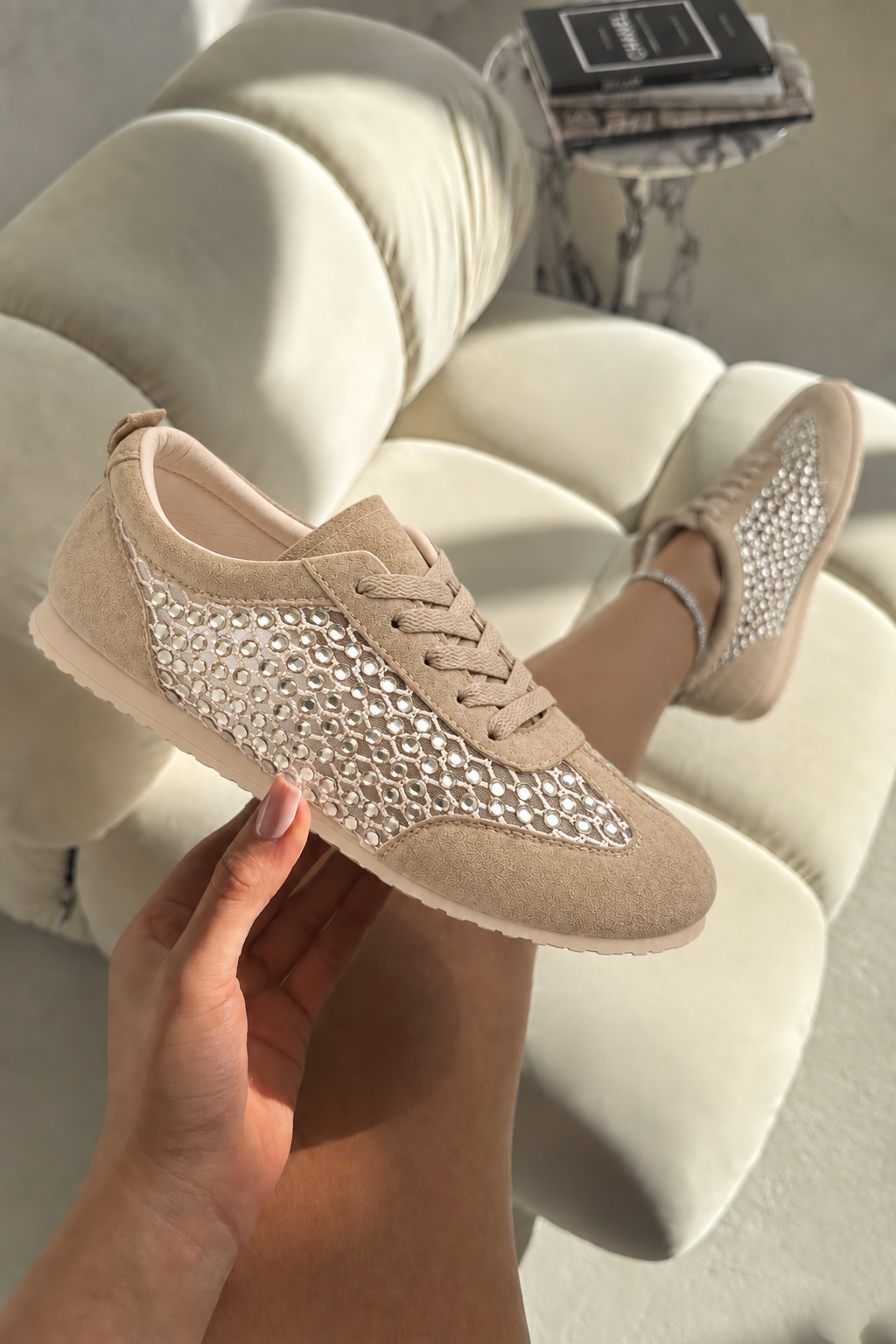 A pair of beige low-top sneakers with a textured rhinestone-embellished side panel, shown on a person&