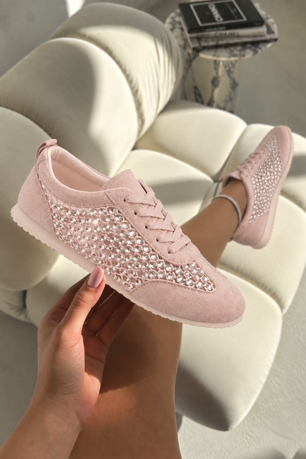 A pair of light pink low-top sneakers with sparkling rhinestone embellishments across the sides, shown with one shoe being held by a hand and the other worn on a foot, set against a modern white tufted sofa.