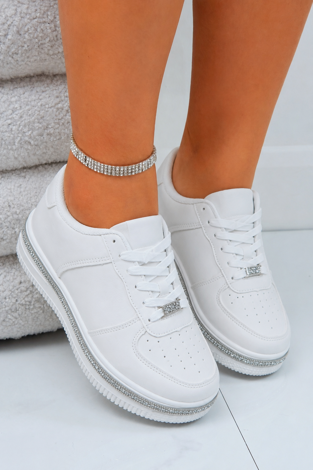 A pair of white low-top sneakers with silver rhinestone trim along the soles and lace accents, shown on a model&
