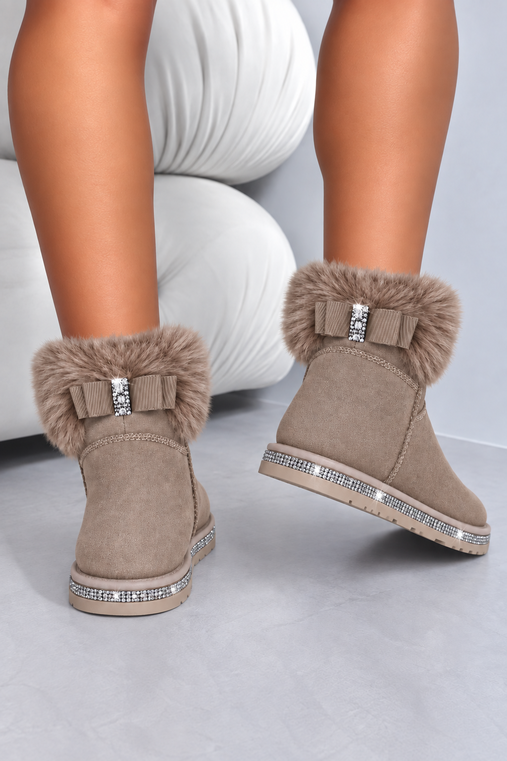 Taupe faux-suede ankle boots with fluffy faux-fur trim, decorative bows, and rhinestone embellishments, shown on a model&