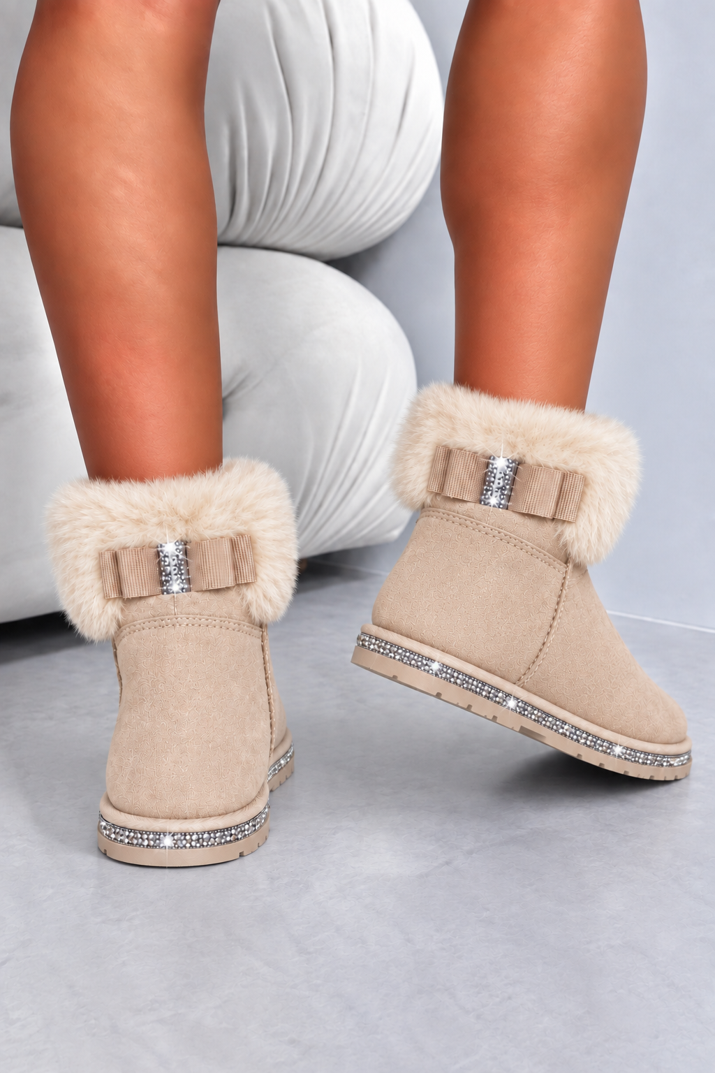 Beige suede ankle boots with fluffy cream faux-fur trim, decorative bows with rhinestone centers, and crystal-studded soles, shown on a model&