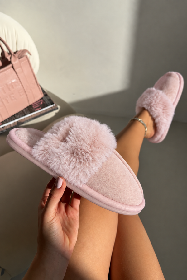 A pair of light pink fuzzy slip-on slippers, with one being held by a hand and the other worn on a foot, in a brightly lit indoor setting.