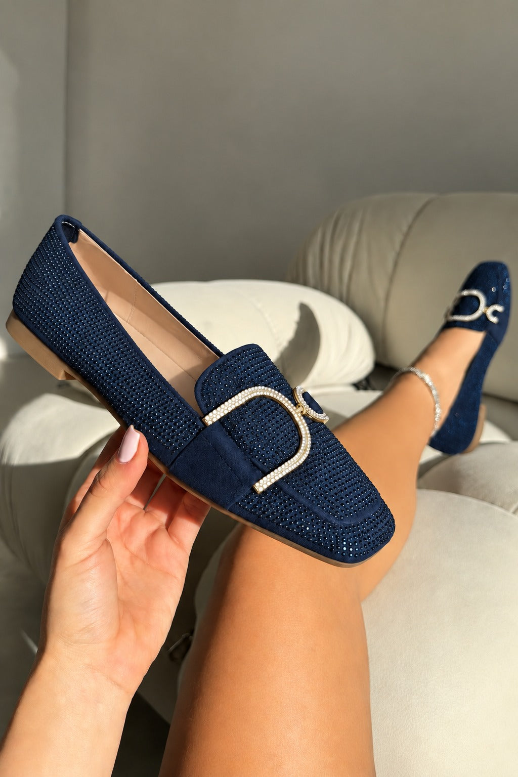 A navy blue woven loafer shoe with a silver-tone horsebit buckle, held in a hand with the other shoe worn on a foot, set against a neutral gray sofa background.
