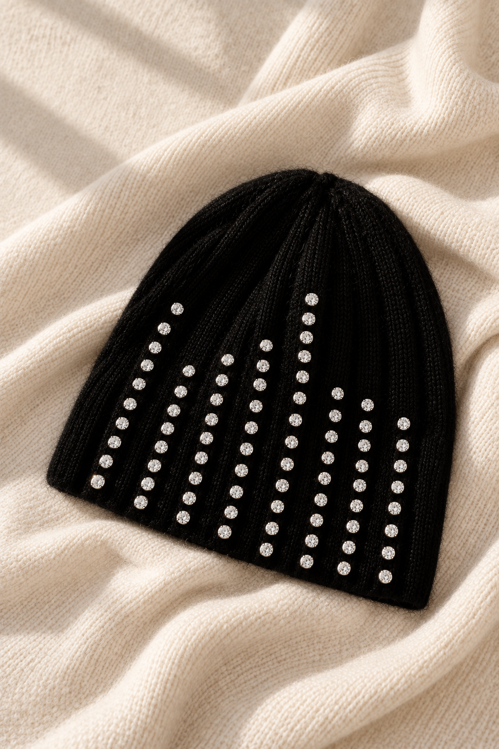 A black knit beanie hat embellished with vertical rows of silver rhinestones, laid flat on a cream-colored textured knit blanket.