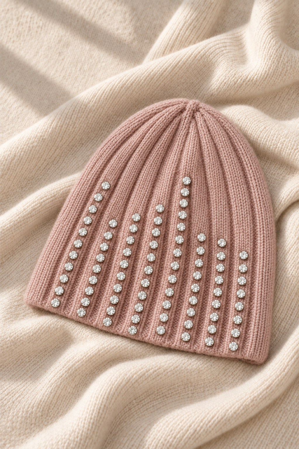 A dusty pink ribbed knit beanie hat embellished with vertical rows of clear rhinestones, laid flat on a cream-colored textured knit blanket.