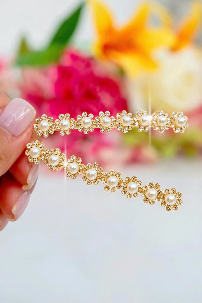 Two gold floral hair clips with white faux pearls, held by a hand against a blurred background of pink and yellow flowers.