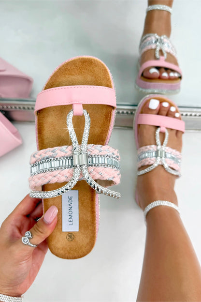 Pink sandals with a bow and rhinestones held by a hand, with another sandal worn on a foot in the background.