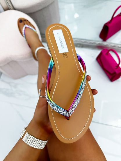 Multicolored sandal with rhinestone details held by a hand, with a blurred background of pink handbag and white shoes.