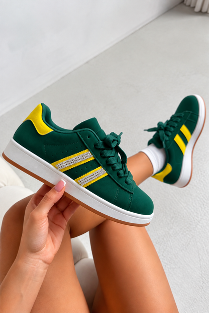 A pair of green low-top sneakers with yellow accents and white soles, shown being worn with one shoe held up by a hand. The background is a clean, light gray indoor floor.
