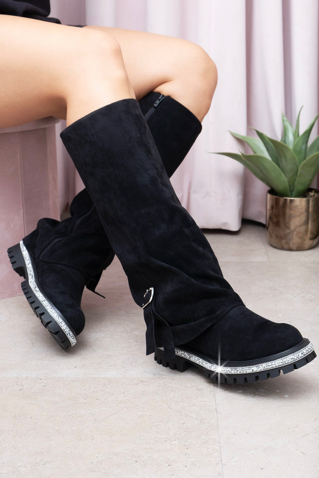 Black knee-high boots with decorative elements worn by a person sitting on a light-colored floor.