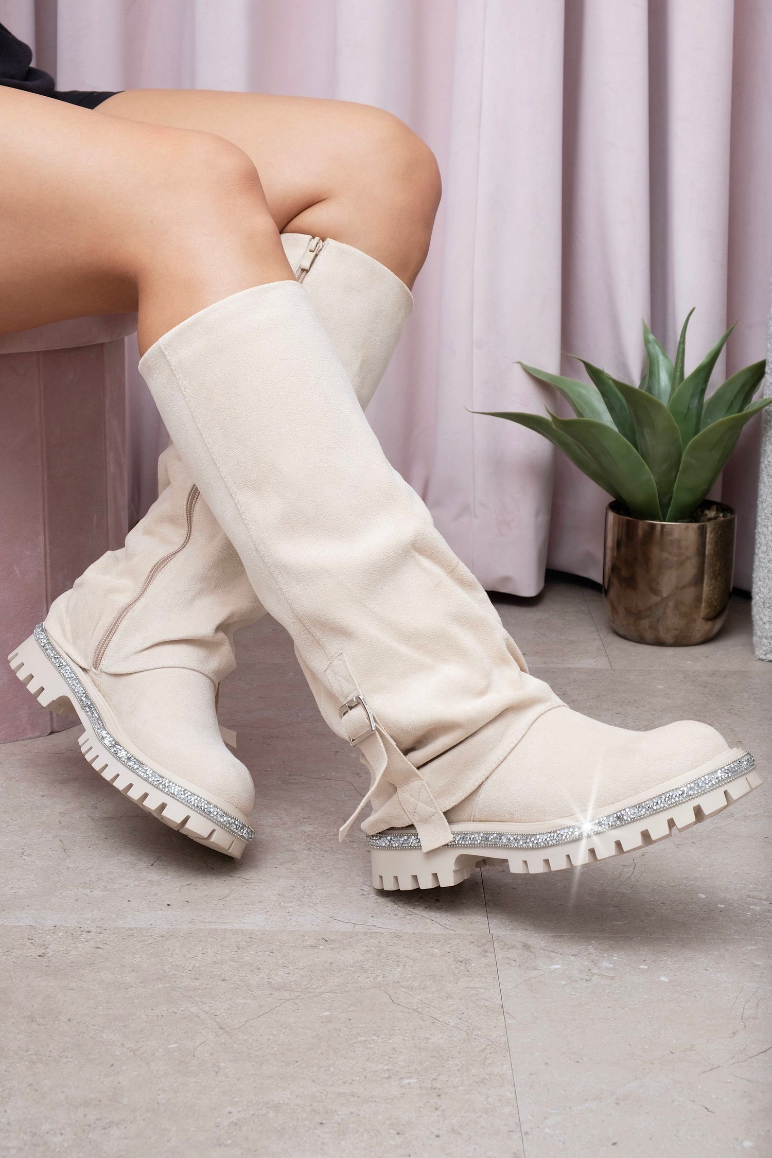 Nude knee-high boots worn by a person on a light wooden floor with a plant in the background.