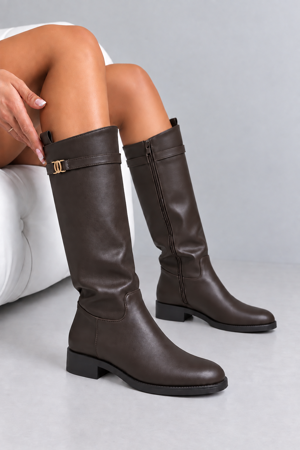 A pair of dark brown knee-high leather boots with a side zipper and a decorative strap detail with a gold-tone buckle, shown on a model sitting on a white tufted chair against a light gray background.