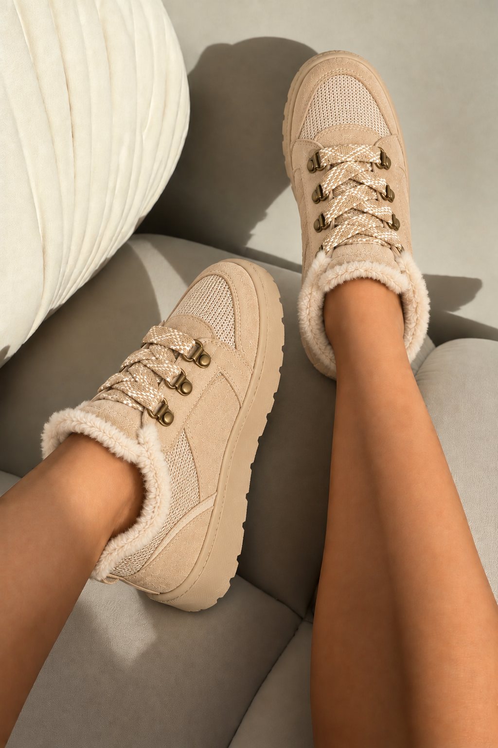 A pair of beige low-top sneakers with fuzzy sherpa lining and gold-tone lace hardware, shown being worn on a person&