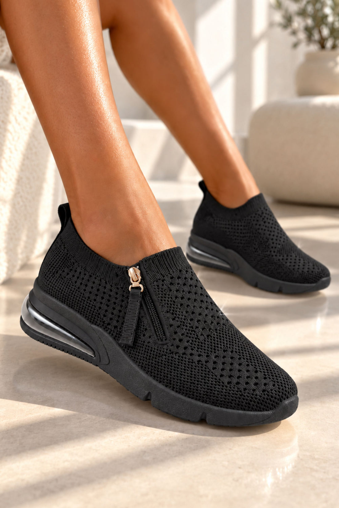 A person wearing black slip-on knit sneakers with a side zipper and tassel detail, shown in a bright indoor setting with sunlight and a light stone floor.